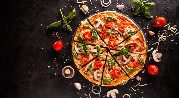 Krojona pizza z pomidorami, pieczarkami i bazylią na czarnym tle, otoczona składnikami.