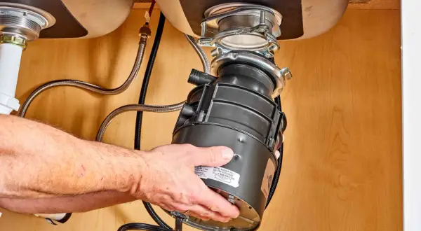 Hands guide a new garbage disposal into place under a sink, part of a garbage disposal installation.