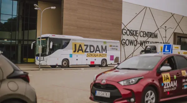 Autobus i czerwony samochód z napisem "JAZDAM SZKOŁA JAZDY" przed budynkiem GOSKIE CENTRUM GOWO-WYSTAWIENNICZE.