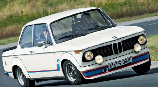 Białe BMW 2002 Turbo z kolorowymi pasami na zderzaku i błotnikach, pędzi po torze.