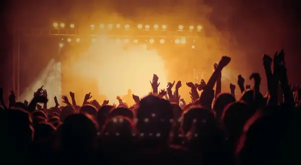 Tłum ludzi z rękami w górze na koncercie, oświetlona scena w tle.