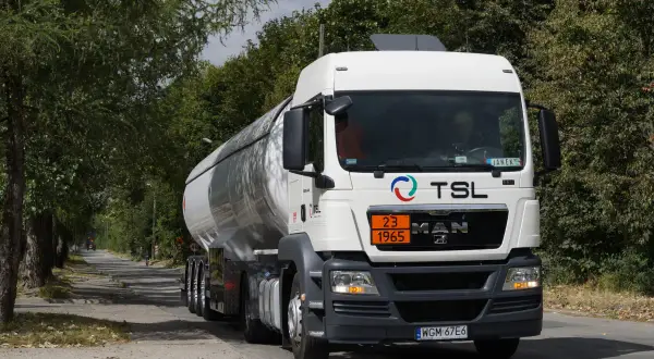 Biały cysterna z logo TSL jedzie drogą wśród drzew. To ważny element branży TSL, transportującej towary.