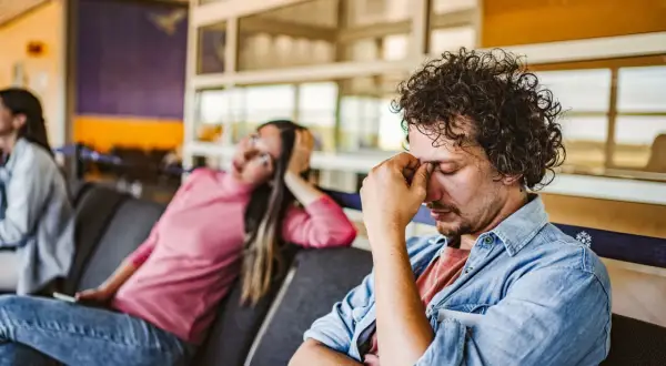 Erschöpfter Mann im Wartebereich, der sich die Augen reibt. Die Frau neben ihm wirkt ebenfalls müde. Gute **Jetlag Tipps** sind hier gefragt.