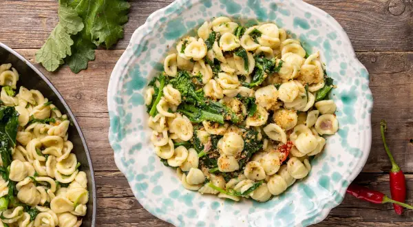 Orecchiette con cime di rapa, la ricetta originale pugliese, servite in un piatto decorato, con peperoncini rossi e foglie di rapa sullo sfondo.