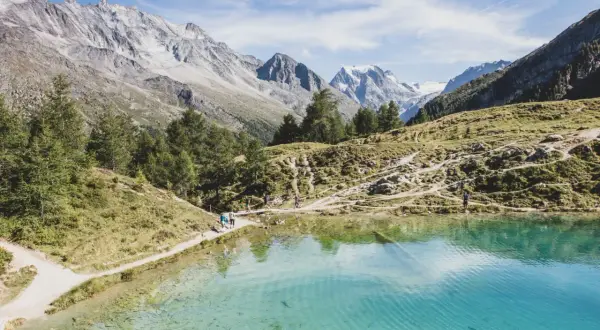 Sentier de randonnée longeant un lac turquoise dans le Val d'Hérens, avec des montagnes majestueuses en arrière-plan.