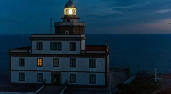 Faro de Finisterre iluminado al anochecer, con el mar oscuro de fondo. Una luz cálida brilla en una ventana.