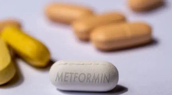 Eine weiße Tablette mit der Aufschrift "Metformin" liegt im Vordergrund. Im Hintergrund sind unscharfe gelbe und orangefarbene Pillen zu sehen. Forschung untersucht, ob Metformin bei Demenz helfen kann.
