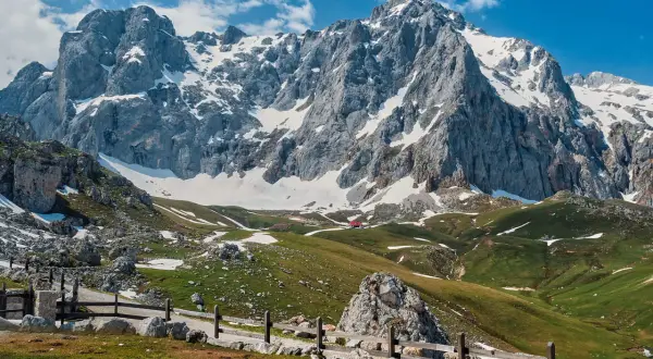 Majestuosas montañas nevadas en los Picos de Europa, cerca de Llanes. Un camino de piedra y madera serpentea por prados verdes salpicados de nieve.