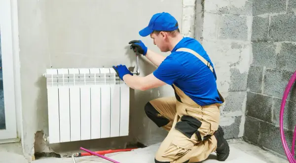 Instalator w niebieskiej czapce i ogrodniczkach pracuje przy grzejniku. Pokazuje, jak zdjąć grzejnik, używając klucza.