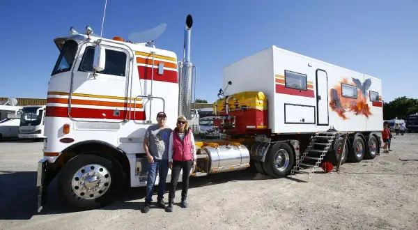 Un couple pose devant un camion américain customisé, avec une remorque aménagée en camping-car ornée d'un aigle et d'un paysage désertique.