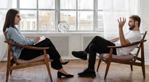 Uomo e donna seduti su sedie di legno, in conversazione. La donna scrive su un quaderno. La prima seduta psicologo si paga, un investimento per il benessere.