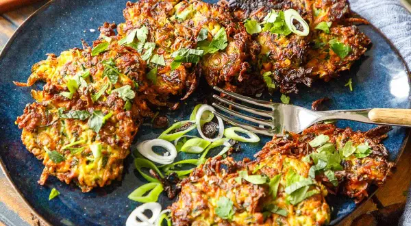 Knusprige Zucchini-Puffer, garniert mit frischen Kräutern und Frühlingszwiebeln, liegen auf einem blauen Teller. Eine Gabel liegt daneben.
