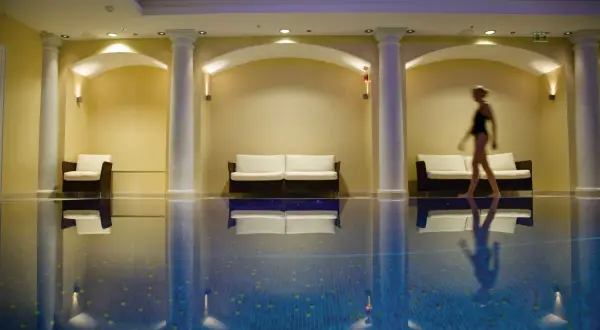 Una mujer camina junto a una piscina interior, rodeada de sofás y columnas, creando una atmósfera de relajación para una experiencia spa sensorial.