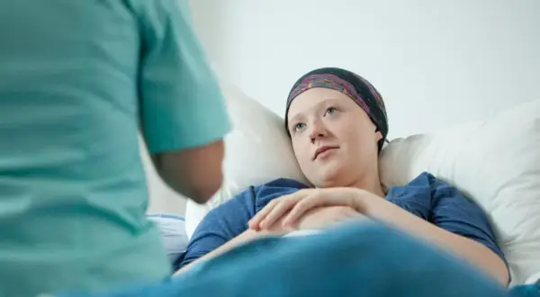Młoda pacjentka w chustce na głowie leży w łóżku, słuchając pielęgniarki. To może być początek walki z chorobą, gdzie rak przedinwazyjny co to znaczy, jest kluczowym pytaniem.