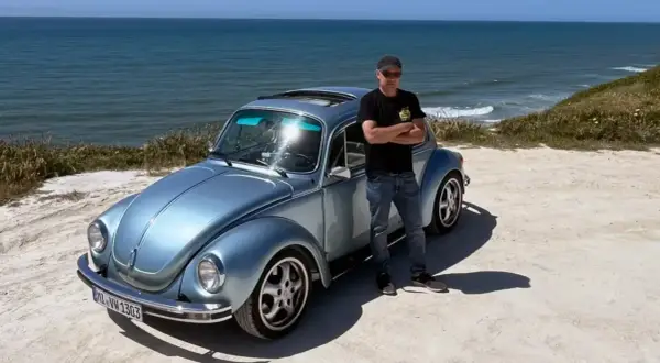 Man står bredvid en ljusblå Volkswagen Bubbla vid havet. En påminnelse om vem grundade Volkswagen och dess ikoniska design.