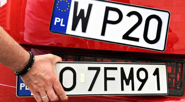 Ręka z bransoletką montuje tablicę rejestracyjną z napisem "O 7FM91" na czerwonym samochodzie. Obok widać inną tablicę "W P20".