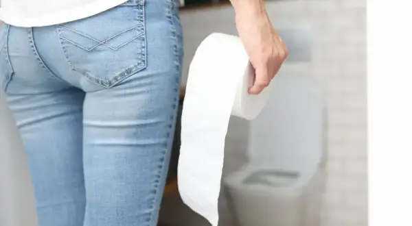 Osoba w dżinsach trzyma rolkę papieru toaletowego, stojąc przed toaletą. Może to sugerować problemy z trawieniem, takie jak biegunka tłuszczowa.
