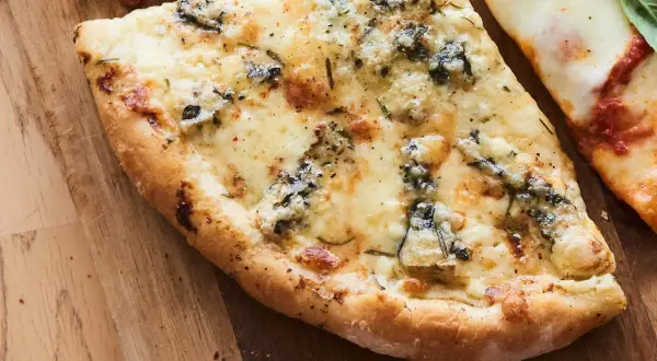 Une part de pizza 4 fromages, avec sa croûte dorée et son mélange de fromages fondus, sur une planche en bois.