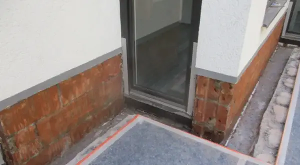 Rote Ziegelsteine am Sockel zeigen Anzeichen von aufsteigender Feuchtigkeit im Sockelbereich. Ein Fensterrahmen ist sichtbar.