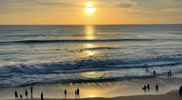 Zachód słońca na plażach Bali. Surferzy czekają na fale, a ludzie spacerują po piasku, ciesząc się złotym blaskiem.