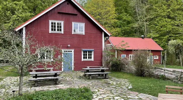 Stocklycke vandrarhem, röda trähus med vita knutar och fönster, omgivna av grönskande skog. Picknickbord och stenlagd uteplats inbjuder till avkoppling.