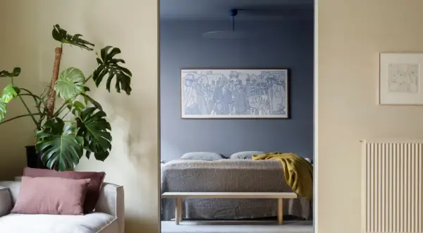 Ett mysigt blått vardagsrum med en stor Monstera och en soffa. Genom en öppning syns ett sovrum med en konstnärlig tavla.