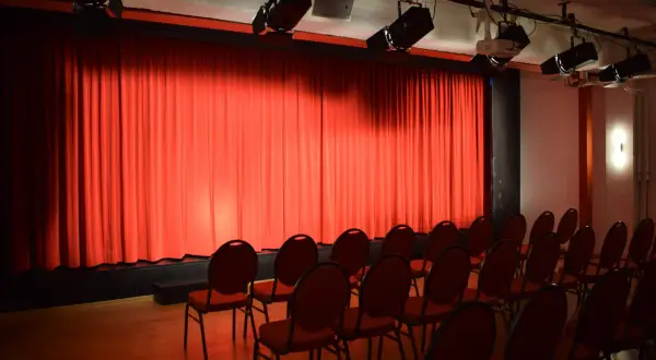 Rote Vorhänge und leere Stühle im **theas theater**. Scheinwerfer sind an der Decke montiert, bereit für die nächste Vorstellung.