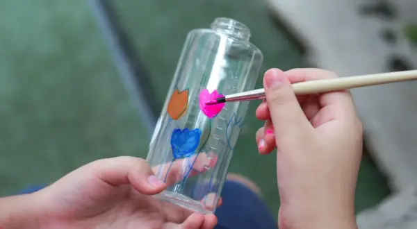 Bambino dipinge fiori su una bottiglia trasparente, un modo creativo per imparare come colorare la plastica trasparente.
