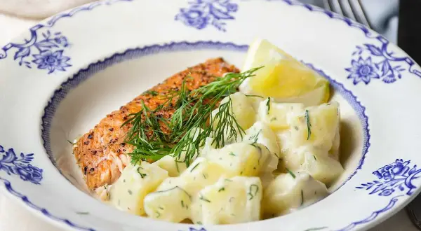 En bit stekt lax med citron och en generös portion krämig dillstuvad potatis på en vacker blåvit tallrik.
