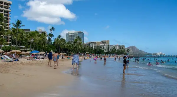Słoneczny dzień na plaży Waikiki, z turystami kąpiącymi się w oceanie i odpoczywającymi na piasku, w tle palmy i hotele.