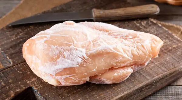 Zamrożona pierś kurczaka na drewnianej desce z nożem w tle.