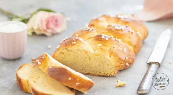 Ein goldbraunes Zopf Brot, bestreut mit Hagelzucker, liegt auf einem Tisch. Daneben eine rosa Rose und eine Tasse.