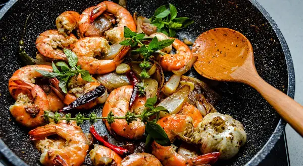 Gambas sautées à l'ail et au piment dans une poêle, prêtes pour le temps de cuisson. Une cuillère en bois est à côté.