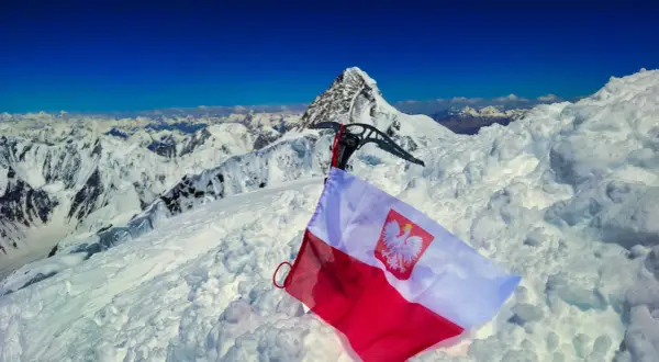 Na śnieżnym szczycie, z lodową siekierą i polską flagą, rozpościera się widok na skalisty, szeroki szczyt i góry.