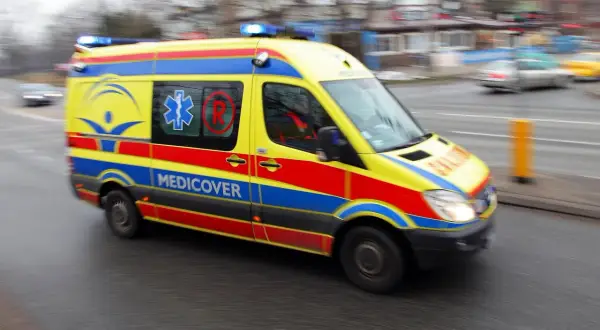 Żółto-czerwono-niebieska karetka Medicover z włączonymi sygnałami świetlnymi, w ruchu. Charakterystyczne oznaczenia karetek na karoserii.