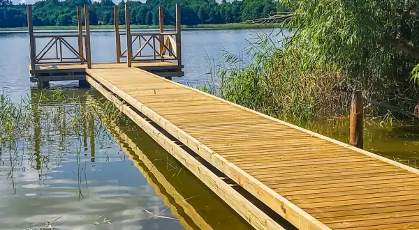 Drewniany pomost nad jeziorem zaprasza do spaceru. Woda odbija błękitne niebo i zielone drzewa.
