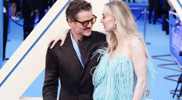 Pedro Pascal y su pareja, con ella luciendo un vestido azul con flecos, se miran tiernamente en una alfombra azul.