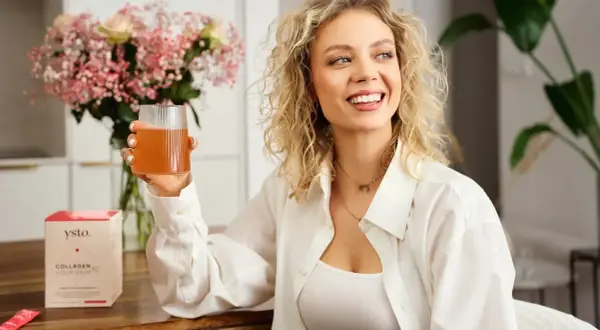 Uśmiechnięta kobieta z kręconymi blond włosami trzyma szklankę napoju. Obok stoi pudełko z napisem "Collagen Your Skin", sugerujące kolagen hydrolizowany.