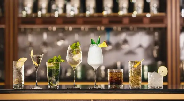 A row of seven colorful cocktails on a bar, with bottles in the background.