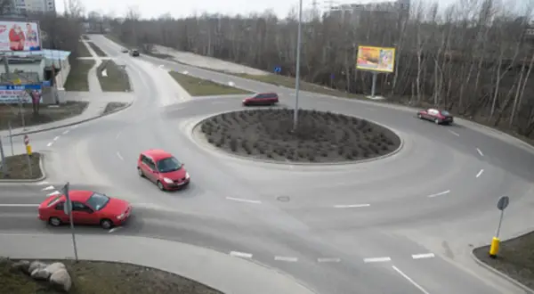 Widok z góry na rondo z kilkoma samochodami, drzewami i billboardami w tle.