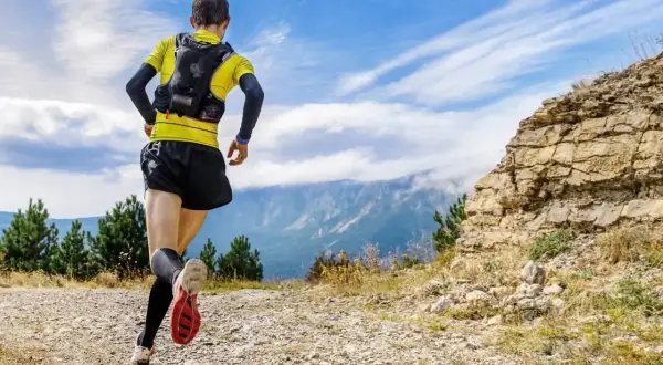 Un coureur en tenue de trail, sac sur le dos, s'élance sur un sentier rocailleux. Le paysage montagneux et le ciel bleu promettent une aventure ultra.