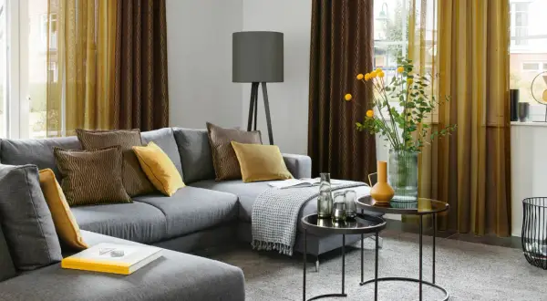 Gemütliches Wohnzimmer mit grauem Sofa, Kissen in Braun und Gelb, sowie moderne Fenstergestaltung mit zweifarbigen Vorhängen.