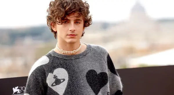 Timothée Chalamet con un ciuffo a tendina uomo, indossa un maglione grigio con cuori e una collana di perle.