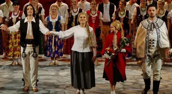 Grupa ludzi w strojach ludowych, mężczyzna w białej koszuli, kamizelce i spodniach z haftem, kobieta w kwiecistej sukni i halka.