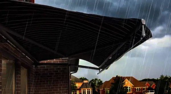A dark, striped awning installation provides shelter from a heavy rain shower over a brick house.