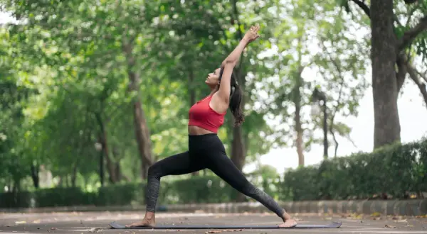 Frau in Krieger-II-Pose, eine von vielen Yoga-Posen, übt im Freien auf einer Matte.