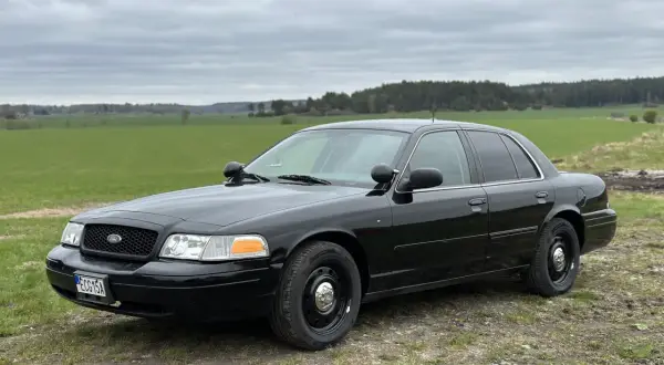 Svart Ford Crown Victoria, en robust bil som står parkerad på en grusväg med gröna fält och skog i bakgrunden.