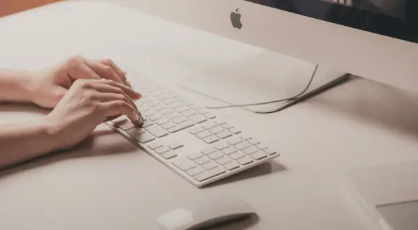 Mani che digitano su tastiera bianca, vicino a un mouse. Attenzione ai sintomi di tendinite da mouse per evitare dolori.