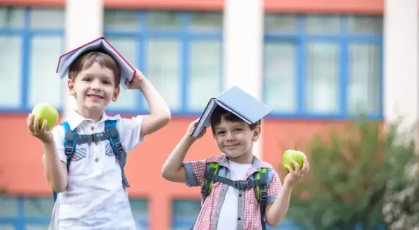 Dwóch chłopców z książkami na głowach i jabłkami w rękach przed szkołą. Zastanawiam się, czy to już 6 klasa, ile lat mają ci mali uczniowie?