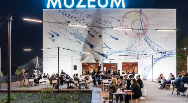Wieczorem muzeum sztuki nowoczesnej nad Wisłą tętni życiem. Ludzie siedzą przy stolikach na zewnątrz, podziwiając abstrakcyjną grafikę na ścianie budynku.